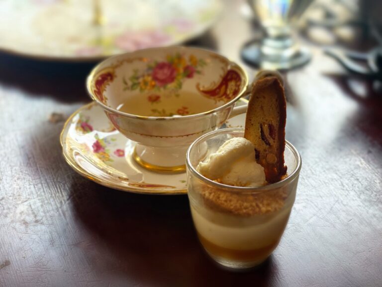Tasse de thé en porcelaine délicate à motifs floraux et liseré doré, accompagnée d'un dessert raffiné sur une table en bois sombre. Reine Vashti