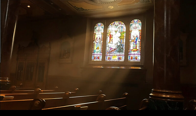 Puissants rayons de soleil traversant la pénombre d'une église depuis un vitrail coloré pour illuminer des bancs en bois verni.