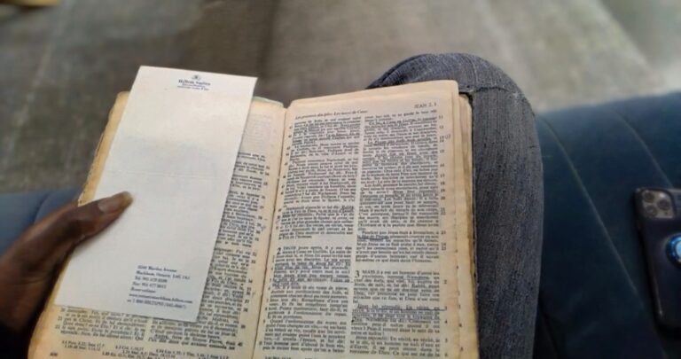 Bible ouverte au chapitre 3 de l'évangile de Jean tenue par une main, posée sur des genoux vêtus de jeans, avec un téléphone à proximité.