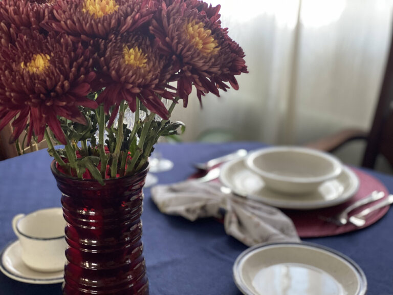 Table soigneusement dressée avec une nappe bleue, de la vaisselle blanche et un bouquet de fleurs rouges, prête pour le repas. Voir Luc 10,38-42.