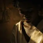 Portrait intimiste d’une femme à la peau foncée, aux traits doux, le regard baissé. Une lumière chaude effleure sa gorge et sa poitrine, révélant un vêtement clair et simple, tandis que le reste de son visage demeure dans l’ombre. L’atmosphère est silencieuse, méditative, comme un moment juste avant la parole. Pourquoi Anokhí choisit l'incarnation à la première personne ? Découvrez comment cette méthode brise le quatrième mur pour une rencontre transformatrice.