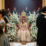 Une jeune femme en robe d'époque assise seule sur un trône doré lors d'un bal majestueux, entourée de couples qui dansent et de compositions florales luxuriantes. Intimité avec Dieu.