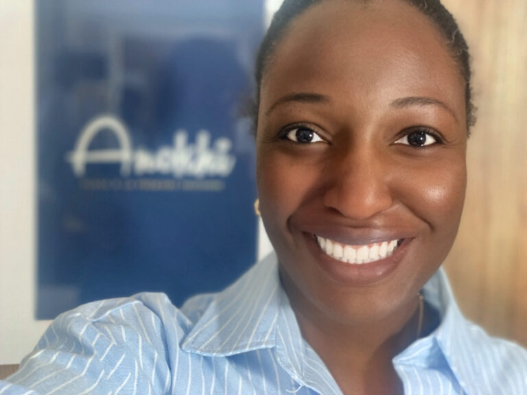 Selfie d'Anathalie souriante au premier plan, vêtue d'une chemise bleue rayée, avec le logo du podcast Anokhí flou en arrière-plan. Anathalie, fondatrice du podcast Anokhí, passionnée de culture hébraïque.