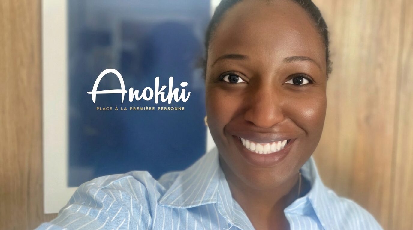 Selfie d'Anathalie souriante au premier plan, vêtue d'une chemise bleue rayée, avec le logo du podcast Anokhí flou en arrière-plan. Anathalie, fondatrice du podcast Anokhí, passionnée de culture hébraïque.