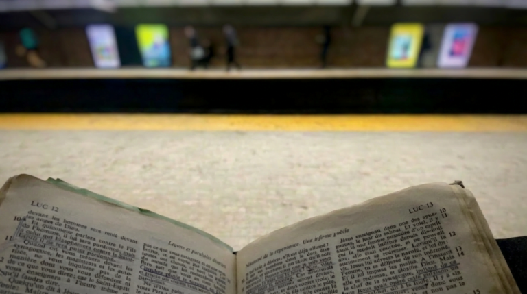 Vue subjective d'une vieille Bible ouverte, posée sur les genoux d'une personne assise sur le quai de la station de métro Mont-Royal à Montréal. Les pages visibles sont Luc 12 et Luc 13, incluant le récit de la guérison de la femme courbée.