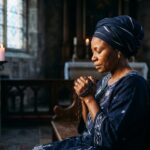 Une femme d'ascendance africaine, vêtue de bleu foncé, prie les yeux fermés dans une église. Elle tient une croix entre ses mains jointes, le visage paisible doucement éclairé par la lueur des bougies en arrière-plan.