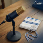 Un microphone professionnel noir et or posé sur une table en bois à côté d'un tallit (châle de prière juif) blanc avec des rayures bleues et dorées. Logo Anokhí en haut à droite.