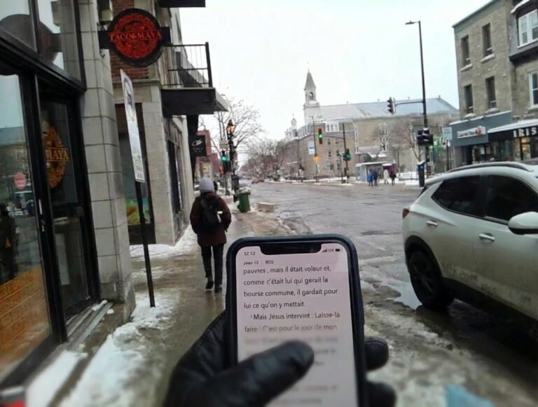 Main gantée tenant un téléphone affichant le texte biblique de Jean 12, avec une rue urbaine enneigée et passante en arrière-plan.