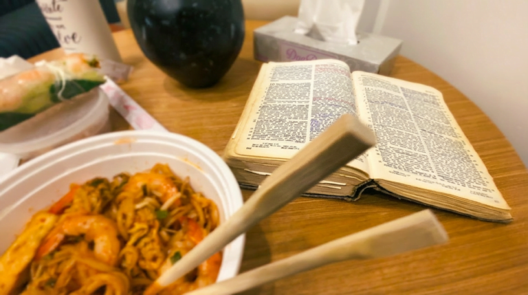 Bol de nouilles et baguettes au premier plan sur une table en bois, avec une Bible ouverte en arrière-plan.